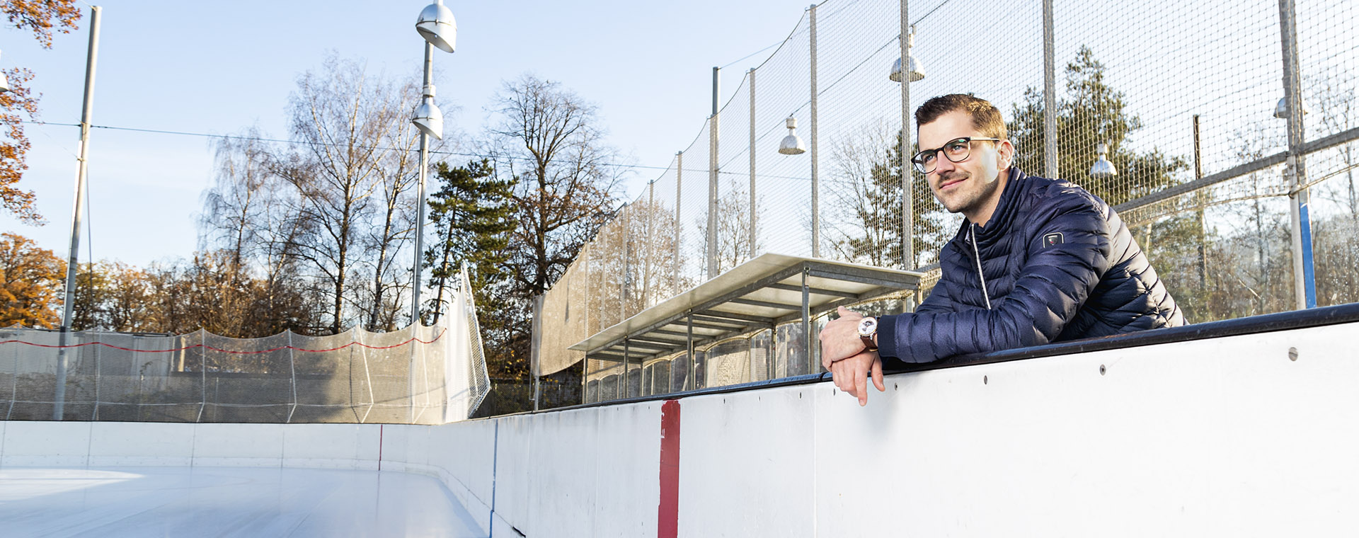 Ein lächelnder Mann mit Brille und einer dunkelblauen Daunenjacke lehnt sich aufmerksam über die Bande eines Eisfeldes oder Sportplatzes und blickt nach rechts in die Ferne. Im Hintergrund sind Bäume ohne Blätter und ein Gitterzaun mit Scheinwerfern zu sehen, was auf eine Sportanlage an einem kühlen, sonnigen Tag hindeutet.