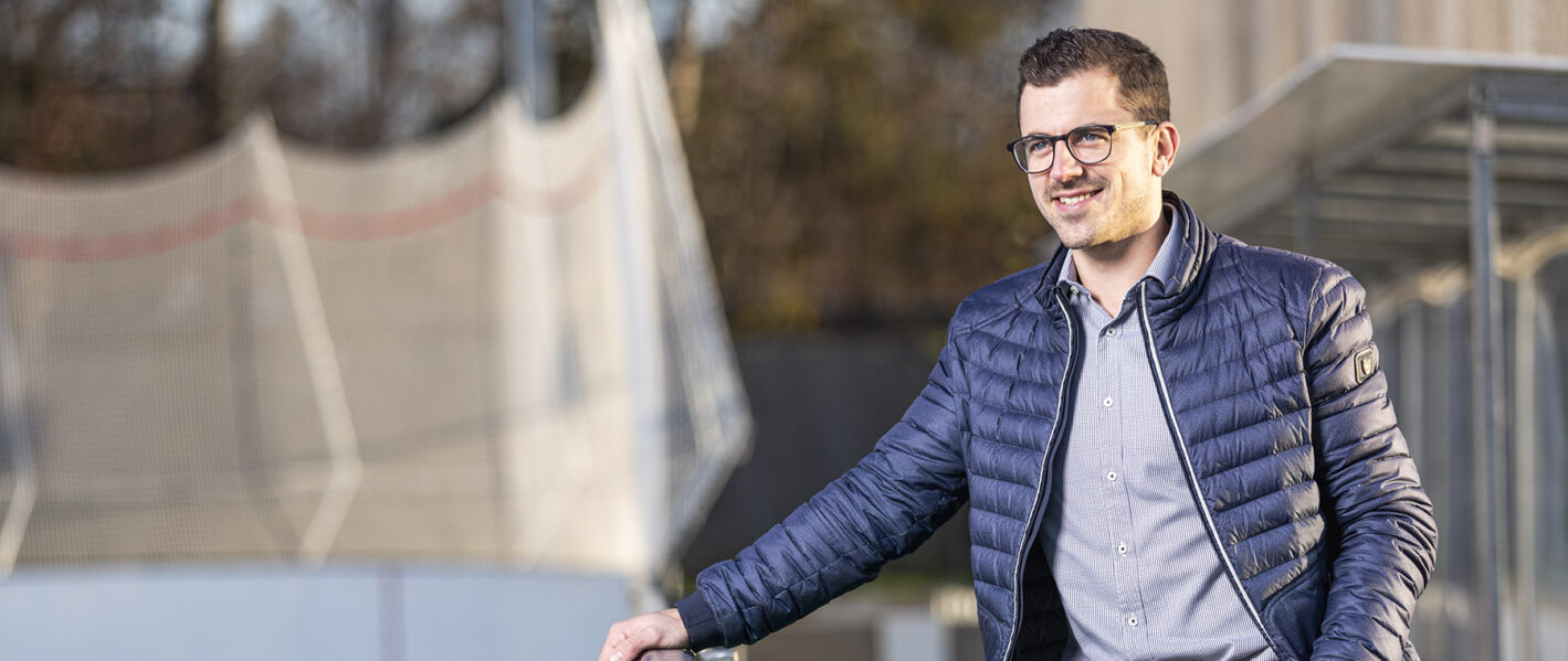 Ein lächelnder Mann mit Brille, einer dunkelblauen gesteppten Jacke und einem gestreiften Hemd blickt zur Seite und lächelt. Er scheint sich auf etwas im Off zu konzentrieren. Im Hintergrund sind unscharfe Gebäude und eine Baustelle oder ein Gerüst zu sehen, was auf eine städtische Umgebung hindeutet.