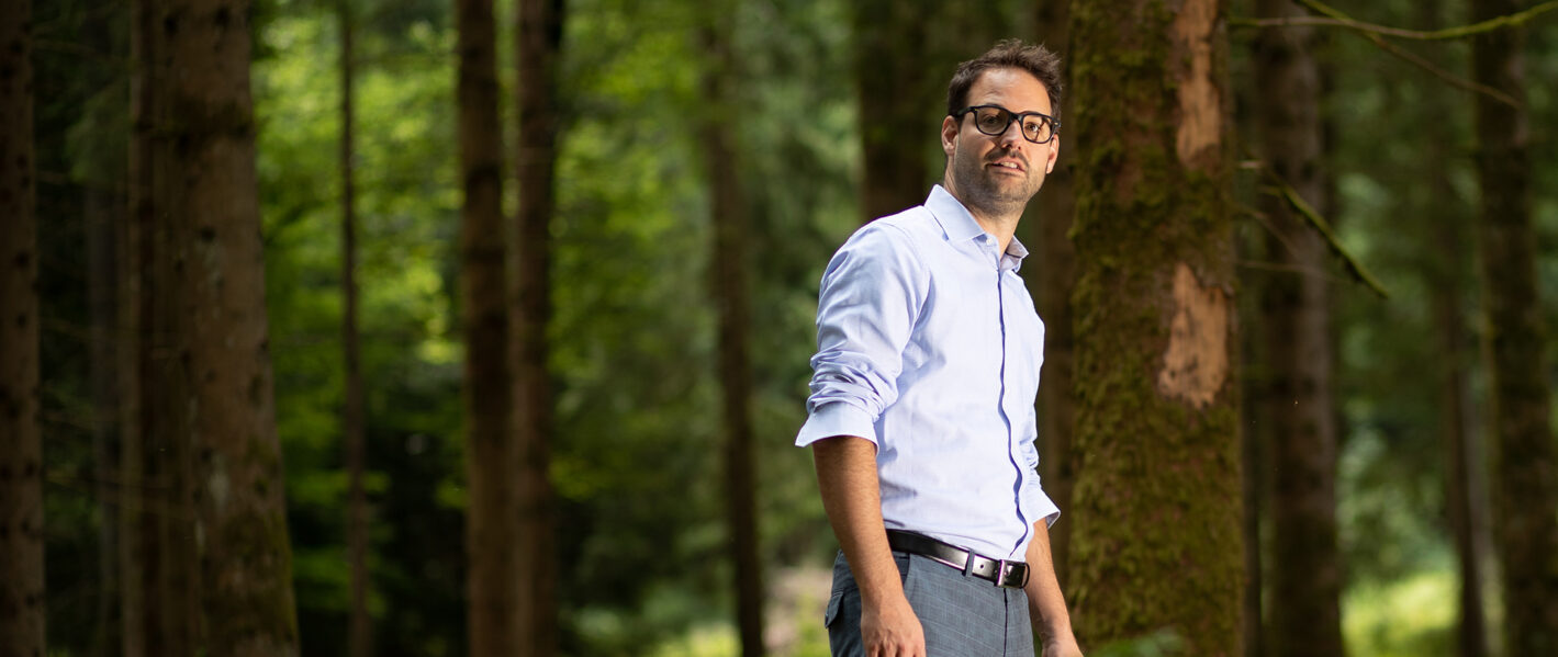 Ein Mann mit Brille und Bart trägt ein hellblaues Hemd und dunkle Hosen. Er blickt nachdenklich und leicht nach oben rechts gerichtet in die Ferne. Im Hintergrund sind hohe, dichte Bäume in einem Wald zu sehen, was eine natürliche und ruhige Atmosphäre schafft.
