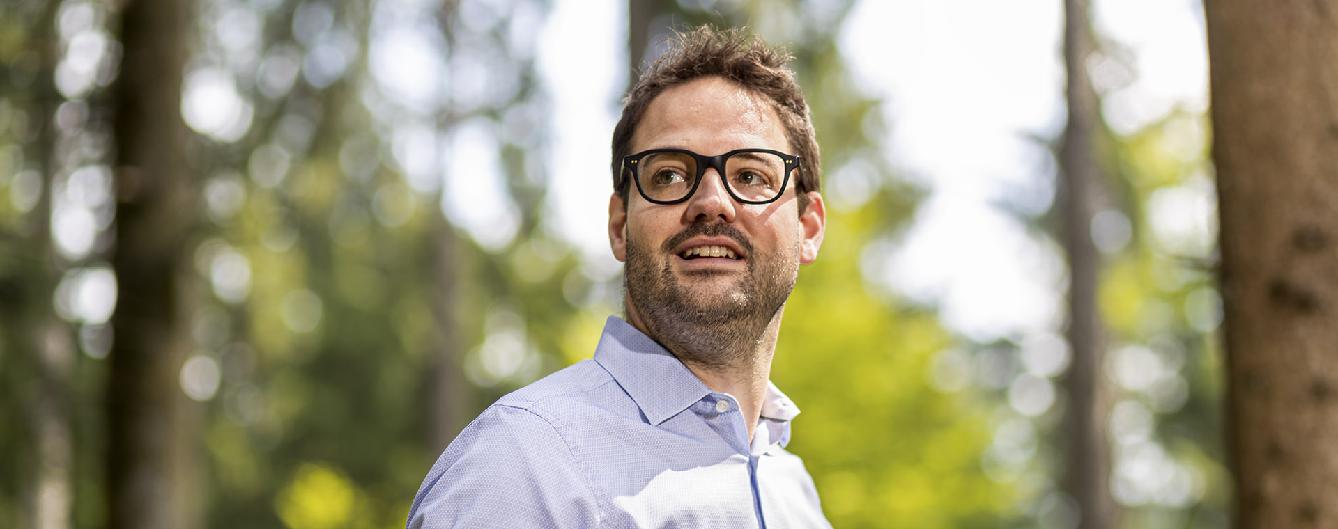 Ein lächelnder Mann mit Bart und dunkler Brille blickt nachdenklich oder interessiert nach oben rechts, sein Mund ist leicht geöffnet. Er trägt ein hellblaues Hemd, und der Hintergrund ist unscharf mit grünem Laub und hellem Sonnenlicht. Das Bild vermittelt einen natürlichen und entspannten Eindruck, als wäre es im Freien aufgenommen.