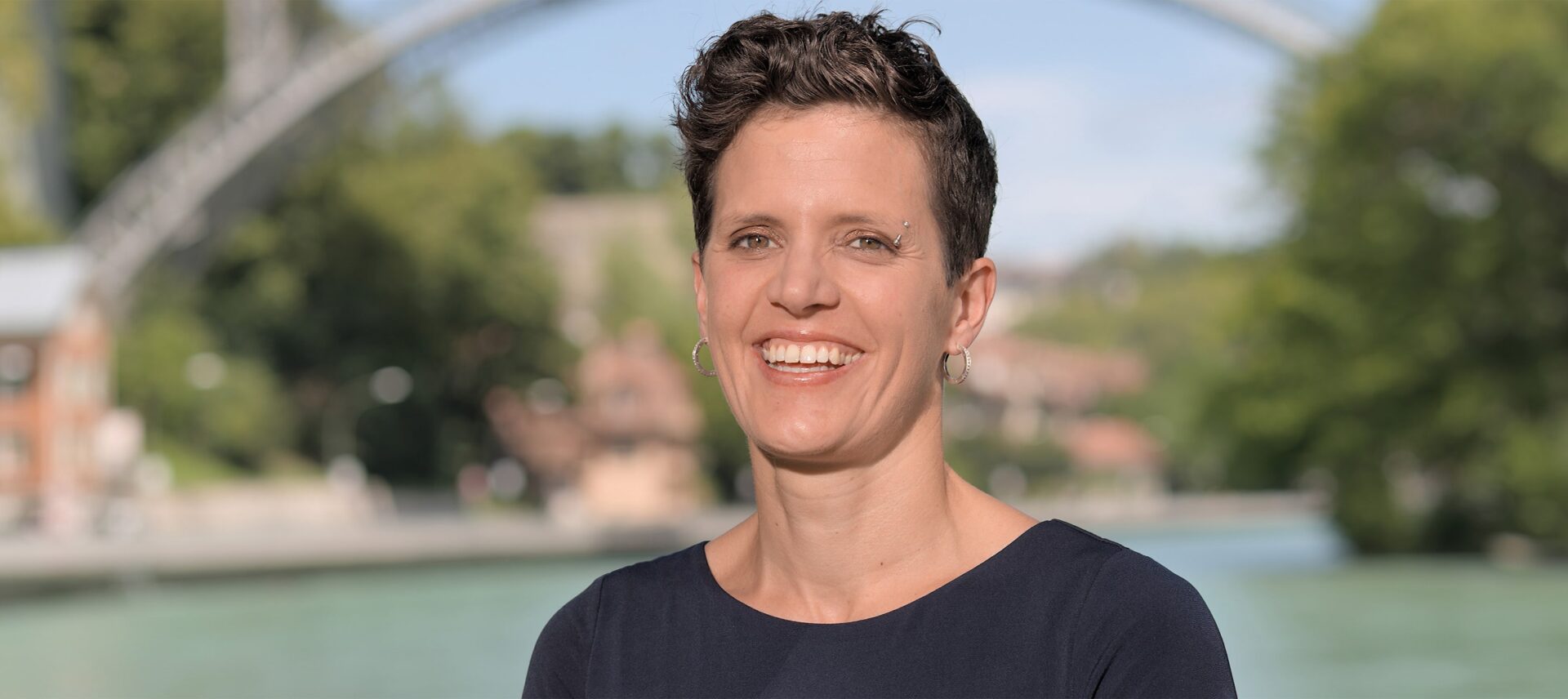 Porträt einer lächelnden Frau mit kurzen Haaren vor einer Flusslandschaft mit Brücke und historischen Gebäuden – offen, sympathisch und professionell.