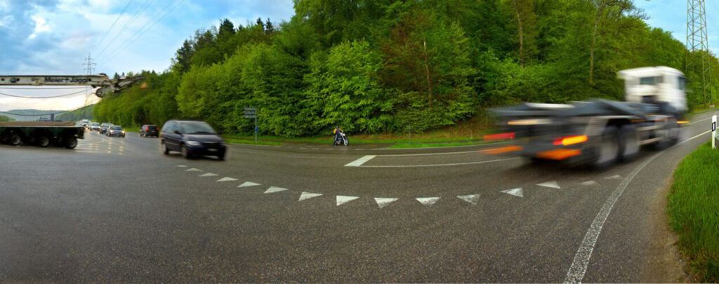Das Panoramabild zeigt eine belebte Strassenkreuzung in einer ländlichen Umgebung. Im Vordergrund biegt ein LKW mit Anhänger, dessen Lichter verschwommen sind, scharf nach rechts ab. Mehrere andere Autos sind in der Ferne auf der Strasse zu sehen. Ein grüner Waldrand säumt die Strasse, und im Hintergrund ist eine Autobahnbrücke oder Überführung zu erkennen. Weisse Dreiecke auf der Fahrbahn markieren den Abbiegebereich.