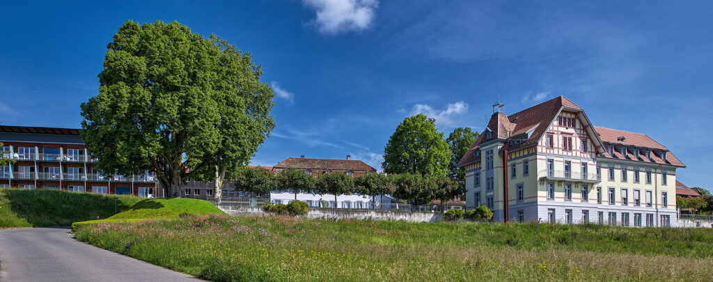 Das Panoramabild zeigt ein malerisches Ensemble von Gebäuden und Bäumen unter einem strahlend blauen Himmel mit einigen weissen Wolken. Im Vordergrund erstreckt sich eine grüne Wiese mit einem Weg, der zu einem historischen Gebäude mit rotem Dach und hellgelber Fassade führt. Dahinter befinden sich weitere Gebäude und üppige grüne Bäume, was auf eine ruhige, ländliche oder vorstädtische Umgebung hindeutet.