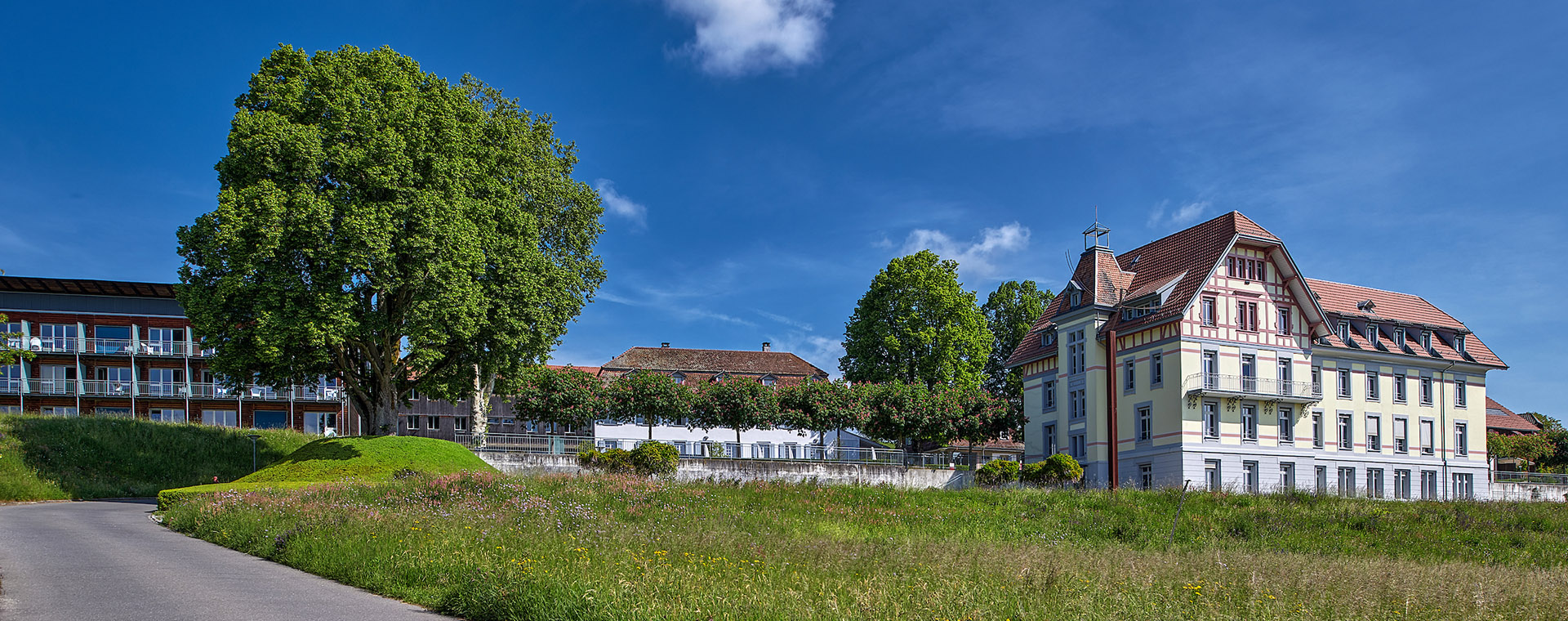 Das Panoramabild zeigt ein malerisches Ensemble von Gebäuden und Bäumen unter einem strahlend blauen Himmel mit einigen weissen Wolken. Im Vordergrund erstreckt sich eine grüne Wiese mit einem Weg, der zu einem historischen Gebäude mit rotem Dach und hellgelber Fassade führt. Dahinter befinden sich weitere Gebäude und üppige grüne Bäume, was auf eine ruhige, ländliche oder vorstädtische Umgebung hindeutet.