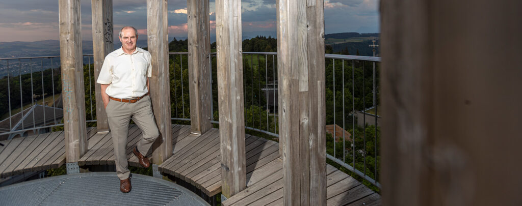 Ein lächelnder Mann in einem weissen Hemd und grauen Hosen steht auf einer Holzplattform eines Aussichtsturms und blickt direkt in die Kamera. Der Turm hat eine hölzerne Struktur mit Säulen und Geländern, die sich um den Betrachter herum erstrecken. Im Hintergrund ist eine weite, grüne Landschaft mit bewaldeten Hügeln und einem dämmrigen Himmel zu sehen, was auf einen Aussichtspunkt in der Natur hindeutet.