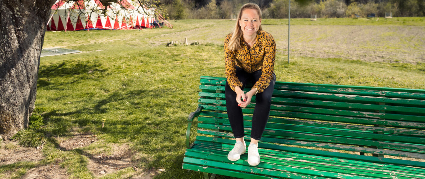 Eine lächelnde, blonde Frau in einem gemusterten, senffarbenen Hemd und dunkler Hose sitzt entspannt auf einer grünen Parkbank im Freien. Im Hintergrund ist eine grosse Rasenfläche mit einem Zirkuszelt oder einem ähnlich gestreiften Bauwerk zu sehen. Die Szene vermittelt eine entspannte und freundliche Atmosphäre an einem sonnigen Tag.