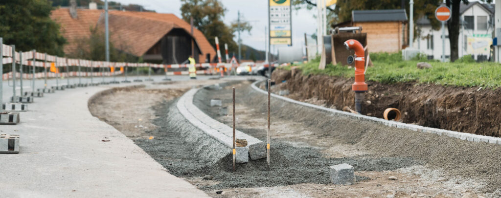 Das Bild zeigt eine Baustelle entlang einer Strasse, auf der neue Gehwege und Rinnen aus grauen Pflastersteinen und Schotter angelegt werden. Links sind Absperrungen und ein fertiger Strassenabschnitt zu sehen, während rechts ein Erdgraben mit Rohren verläuft. Im Hintergrund sind Häuser und Verkehr zu erkennen, was auf Bauarbeiten im ländlichen oder vorstädtischen Raum der Schweiz hinweist.