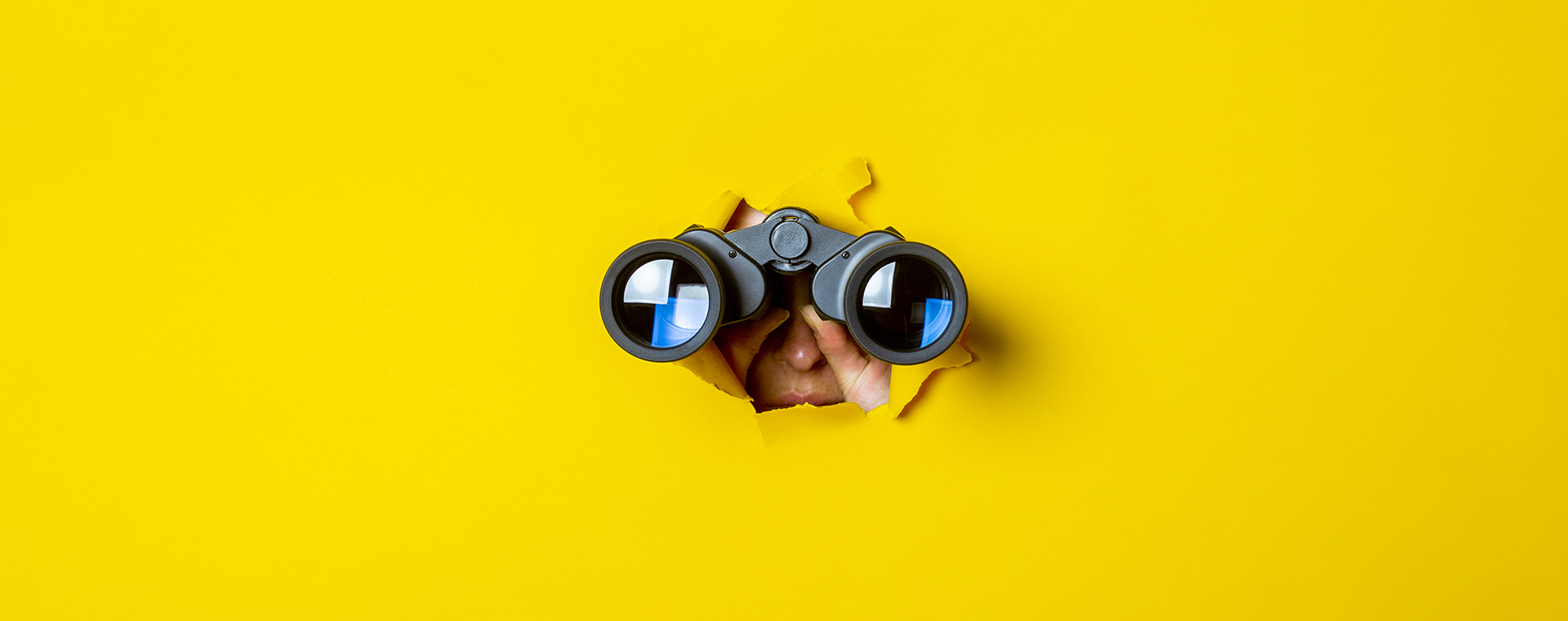 Neugieriger Blick in die Zukunft: Hände halten ein Fernglas durch ein gerissenes Loch in leuchtend gelbem Papier.