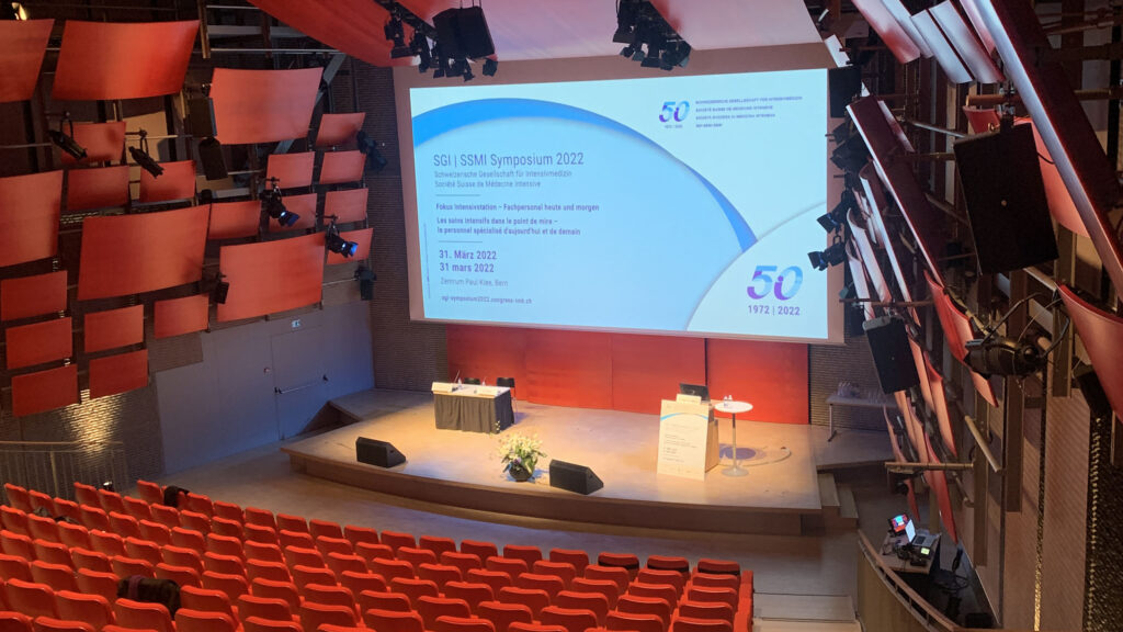 Leerer Hörsaal mit roten Sitzreihen vor einer Bühne mit einer Leinwand für das SGII SSMI Symposium 2022.