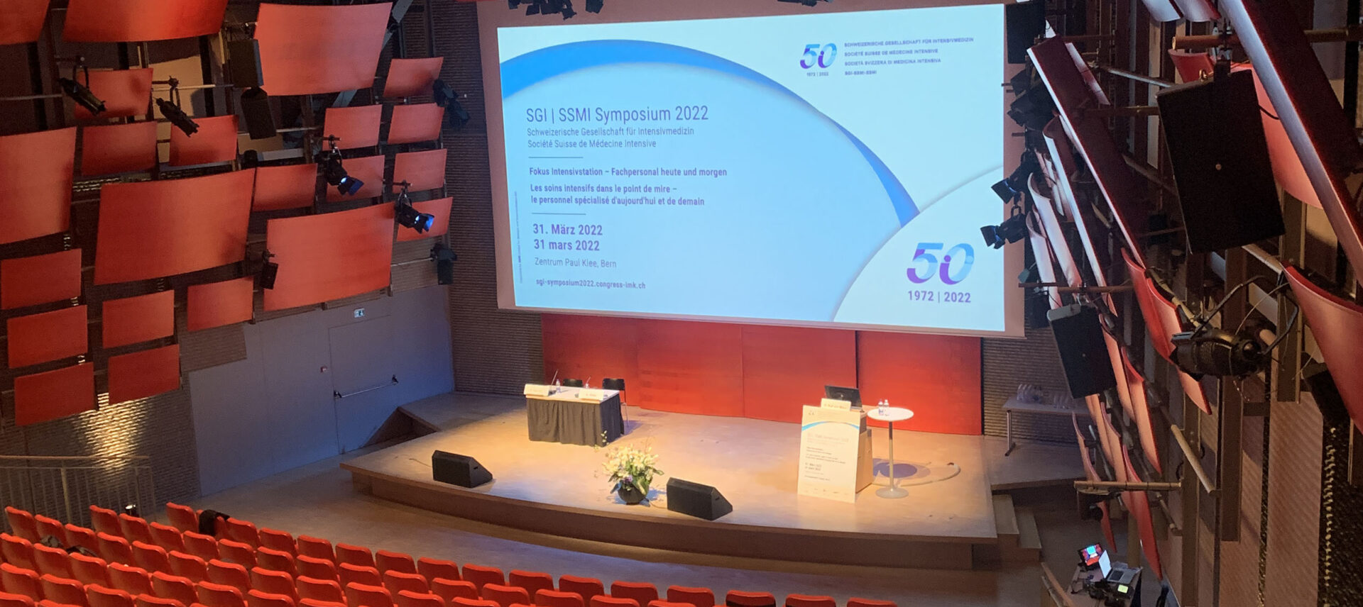 Leerer Hörsaal mit roten Sitzreihen vor einer Bühne mit einer Leinwand für das SGII SSMI Symposium 2022.