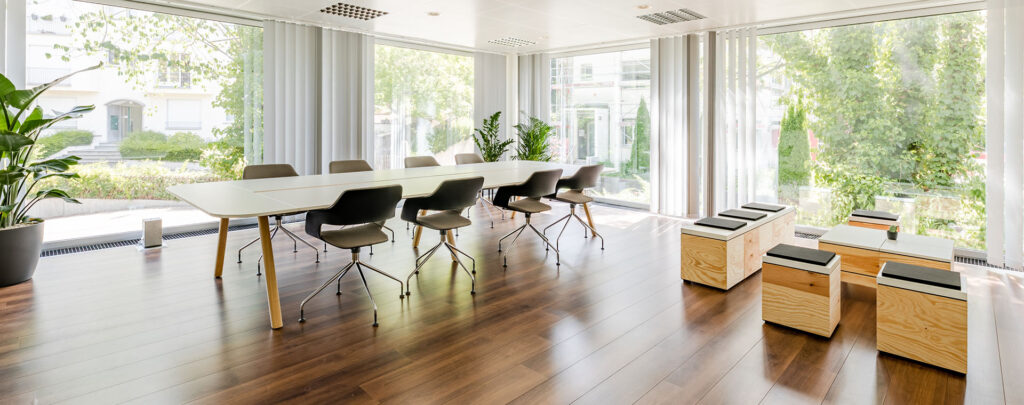 Das Bild zeigt einen hellen und modernen Büroraum mit einem langen, weißen Tisch, um den mehrere graue Bürostühle mit Rollen stehen. Große Fenster lassen viel Tageslicht herein und bieten einen Blick ins Grüne. Der Boden ist aus dunklem Holzparkett.
