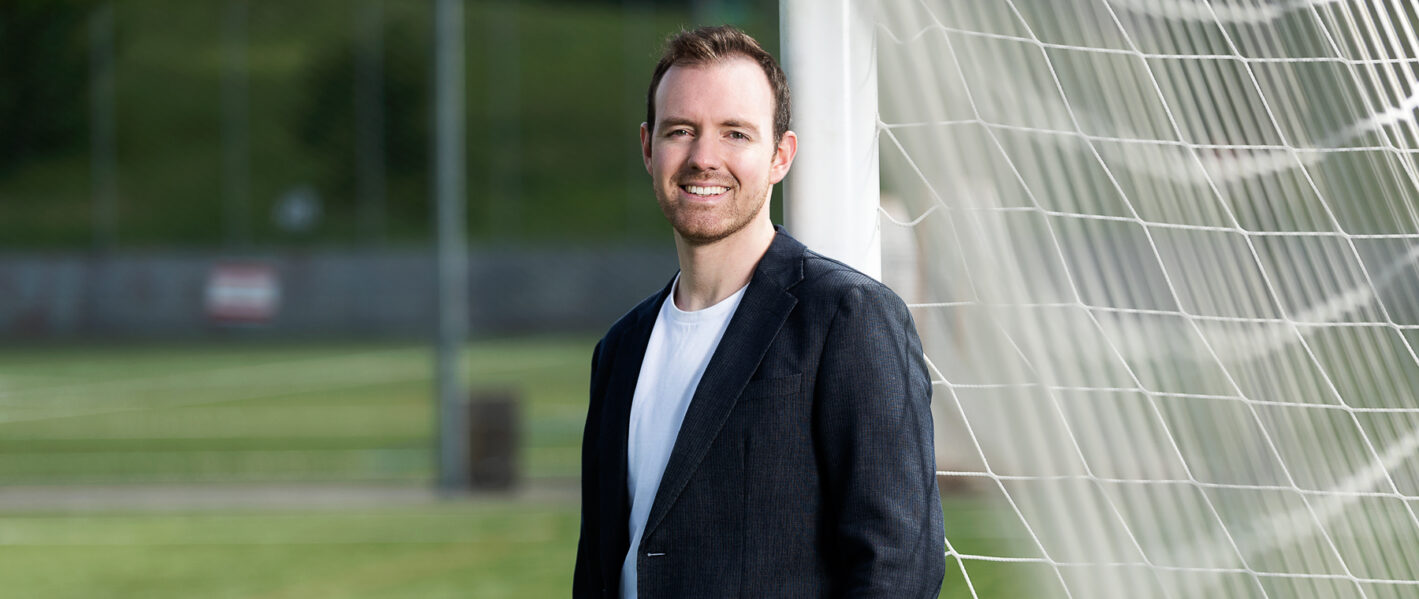 Alt-Text: Ein junger Mann mit braunen Haaren und einem leichten Bart steht lächelnd neben einem weißen Fußballtornetz. Er trägt ein weißes T-Shirt unter einem dunkelblauen Sakko. Der Hintergrund zeigt einen grünen Fußballplatz und unscharfe Gebäude.