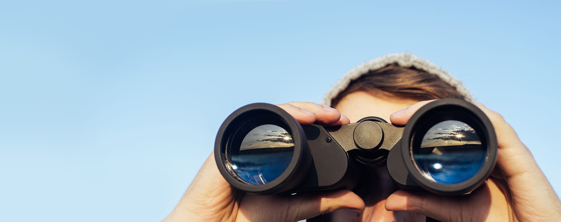 Mann mit Mütze blickt durch Fernglas mit blauem Himmel im Hintergrund.
