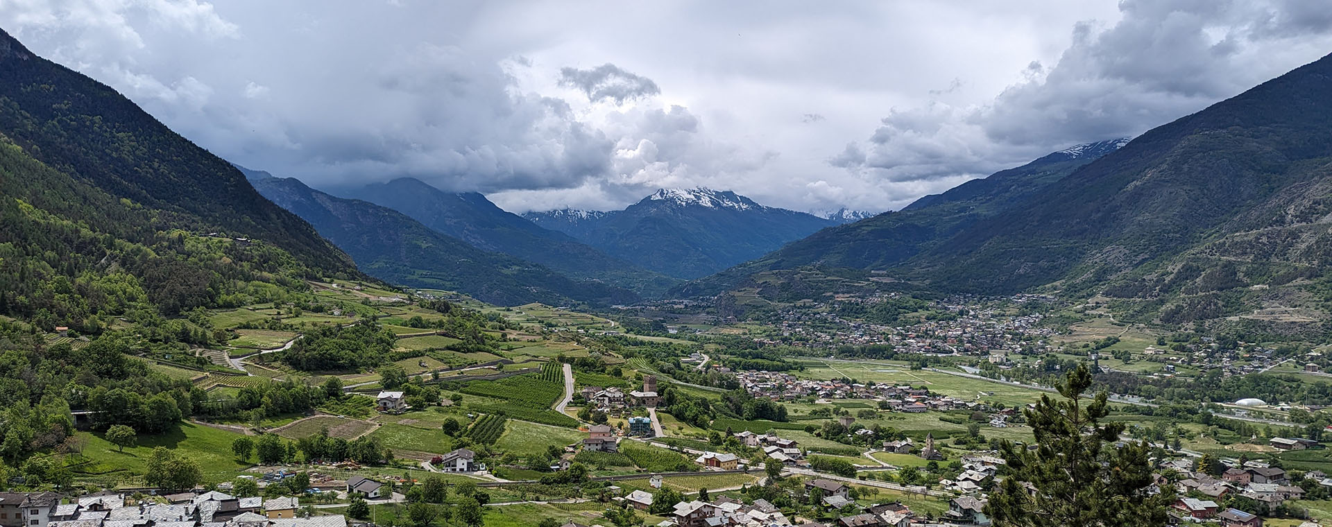 Eine Panoramaaufnahme zeigt ein weites Tal, umgeben von steilen Bergen, deren Gipfel teilweise von Schnee bedeckt sind. Der Himmel ist von grauen Wolken verhangen. Im grünen Tal sind verstreute Siedlungen mit Häusern und Feldern sowie gewundene Straßen sichtbar. Die Landschaft wirkt ruhig und weitläufig.