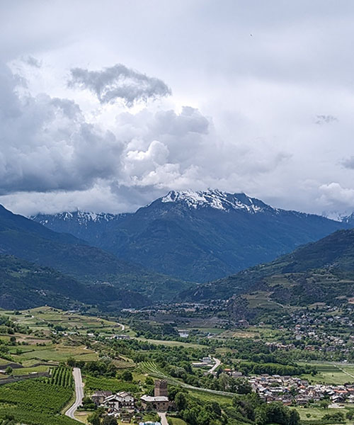 Eine weite Landschaftsaufnahme zeigt ein grünes Tal, eingebettet zwischen hohen, dunkel bewaldeten Bergen, deren Gipfel teilweise mit Schnee bedeckt sind. Der Himmel ist bedeckt mit grauen und weißen Wolken, die über der Szenerie liegen. Im Tal sind kleine Siedlungen mit Häusern und Feldern sowie eine gewundene Straße zu erkennen. Die Perspektive ist von einer erhöhten Position aufgenommen und vermittelt einen Eindruck von der Weite und Erhabenheit der Berglandschaft.