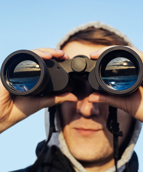 Ein junger Mann mit einer Kapuze über dem Kopf schaut durch ein Fernglas. In den Linsen des Fernglases spiegelt sich eine Landschaft mit Wasser und einem hellen Horizont wider. Der Himmel im Hintergrund ist klar und blau.