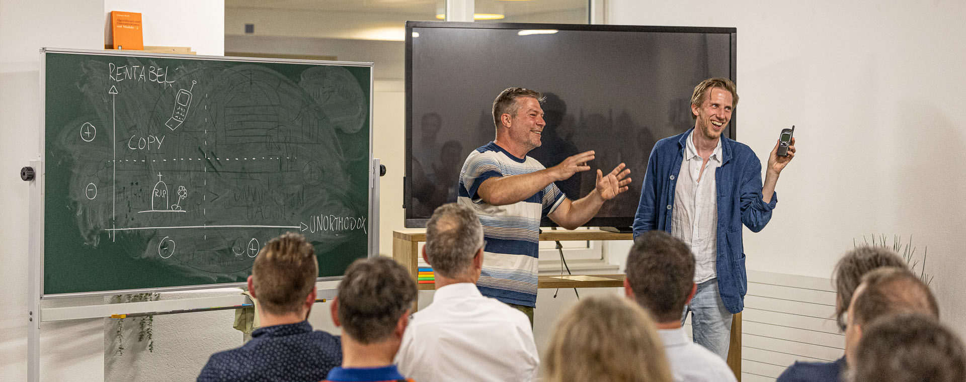 Zwei Männer stehen vor einer Gruppe von Zuhörern in einem Raum in Bern und präsentieren etwas. Der Mann links gestikuliert lebhaft neben einer Tafel mit handschriftlichen Notizen und Diagrammen, während der Mann rechts ein kleines Gerät in die Höhe hält und lächelt. Die Zuhörer sitzen auf Stühlen und blicken auf die beiden Präsentatoren. Im Hintergrund sind ein großer Bildschirm und helle Wände zu sehen.