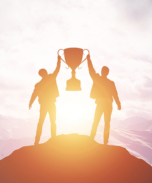 Zwei jubelnde Silhouetten von Personen stehen auf einem Berggipfel und heben gemeinsam eine große goldene Trophäe in die Höhe. Das warme Licht der untergehenden oder aufgehenden Sonne umgibt sie und betont ihren Erfolg. Die Szene vermittelt ein Gefühl von Triumph, Teamwork und dem Erreichen eines gemeinsamen Ziels in einer herausfordernden Umgebung.