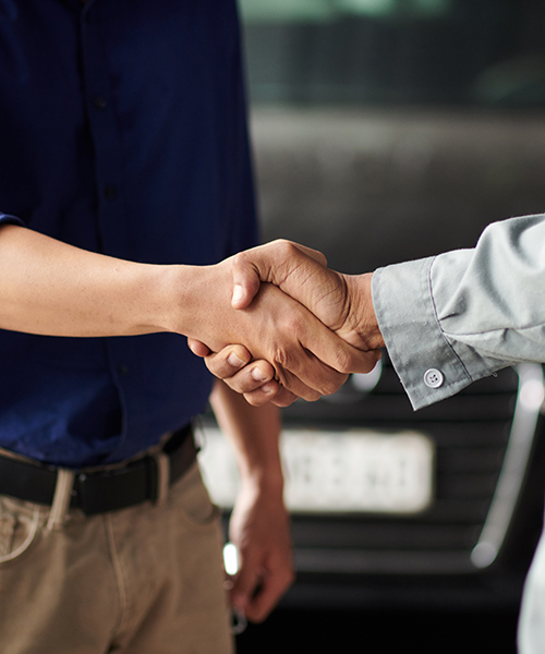 Zwei Personen schütteln sich die Hände vor einem dunklen Hintergrund, der möglicherweise ein Auto oder eine Werkstatt andeutet. Die linke Person trägt ein dunkelblaues Hemd und beige Hosen mit einem schwarzen Gürtel. Die rechte Person trägt eine hellgraue Arbeitskleidung mit hochgekrempelten Ärmeln, an deren Manschette ein Knopf sichtbar ist.
