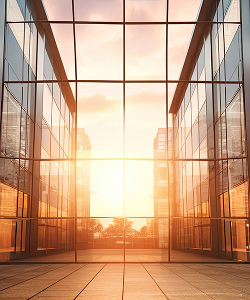 Die Innenansicht eines modernen Gebäudes mit großen Glasfenstern, die das goldene Licht der untergehenden oder aufgehenden Sonne und die Spiegelbilder gegenüberliegender Gebäude einfangen. Der Boden ist mit hellen Fliesen ausgelegt.