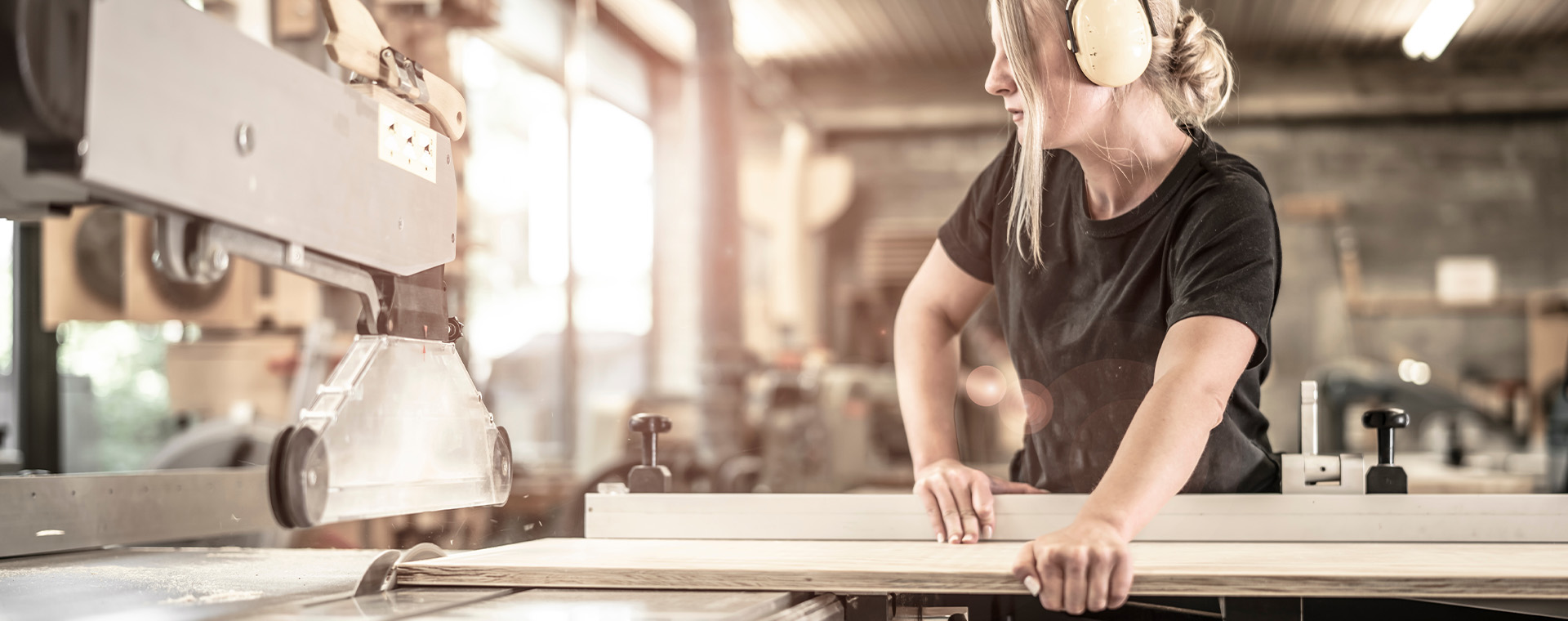 Eine Frau mit blonden Haaren und gelben Gehörschützern arbeitet konzentriert an einer Holzbearbeitungsmaschine. Ihre Hände führen ein langes, helles Holzbrett über eine Säge oder eine ähnliche Werkzeugvorrichtung. Der Hintergrund zeigt eine Werkstattumgebung mit weiteren Maschinen und Holzmaterialien, die auf handwerkliche Tätigkeit hindeuten.