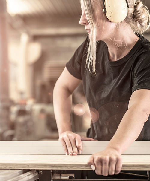Eine Frau mit blonden Haaren und gelben Gehörschützern arbeitet konzentriert an einer Holzbearbeitungsmaschine. Ihre Hände führen ein langes, helles Holzbrett über eine Säge oder eine ähnliche Werkzeugvorrichtung. Der Hintergrund zeigt eine Werkstattumgebung mit weiteren Maschinen und Holzmaterialien, die auf handwerkliche Tätigkeit hindeuten.