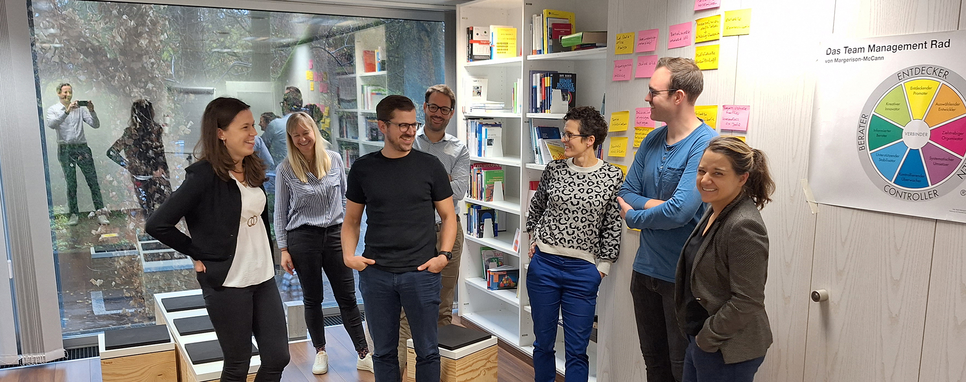 Eine Gruppe von sieben Personen steht in einem hellen Büroraum und scheint sich zu unterhalten oder an einer informellen Besprechung teilzunehmen. Im Hintergrund befinden sich Regale mit Büchern und Ordnern, ein Flipchart mit Notizen und ein Poster mit einem farbigen Diagramm. Durch eine große Fensterfront sind Bäume und schemenhafte Figuren im Außenbereich zu sehen. Die Atmosphäre wirkt entspannt und kollegial.