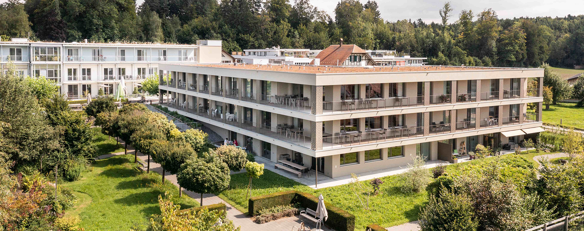 Ansicht eines modernen Mehrfamilienhauses welches als Alters- und Pflegeheim genutzt wird mit Flachdach und Balkonen, umgeben von einem Garten mit Bäumen.