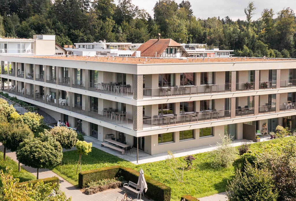 Ansicht eines modernen Mehrfamilienhauses welches als Alters- und Pflegeheim genutzt wird mit Flachdach und Balkonen, umgeben von einem Garten mit Bäumen.