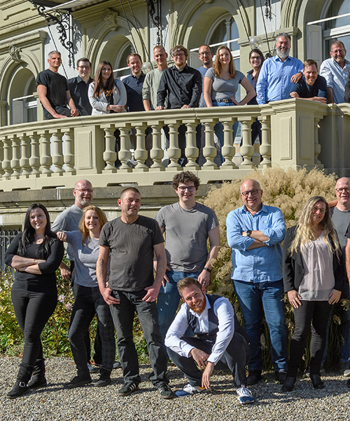 Eine große Gruppe von Männern und Frauen posiert für ein Gruppenfoto vor einem eleganten, hellen Gebäude. Einige stehen auf einem Balkon im ersten Stock, während andere davor auf dem Boden stehen oder hocken.