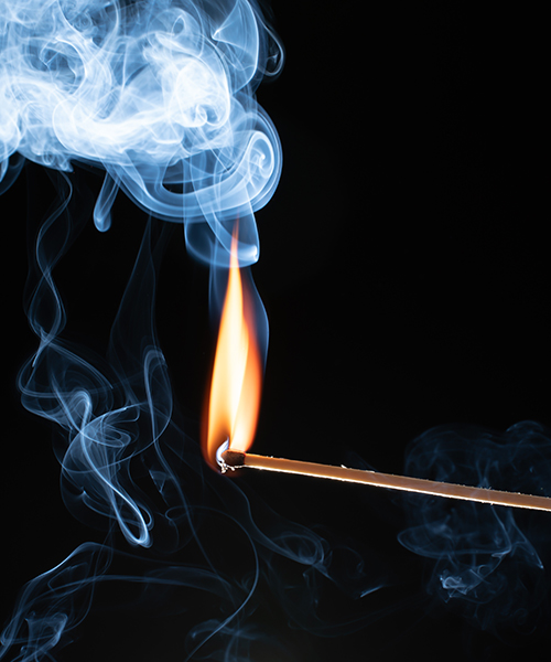 Ein brennendes Streichholz mit einer hellen Flamme und aufsteigendem blau-weißem Rauch vor einem tiefschwarzen Hintergrund.