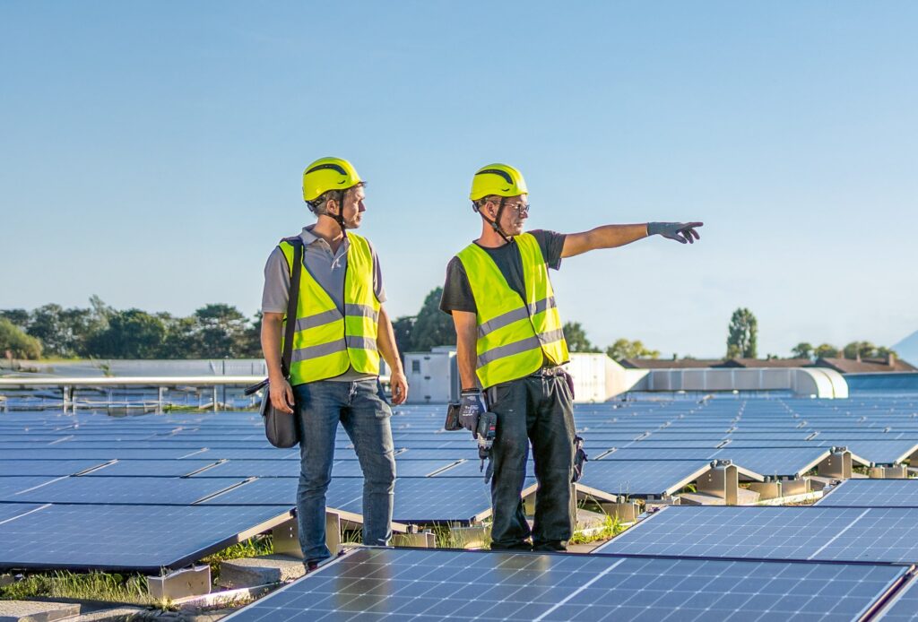 Zwei Arbeiter in Warnwesten und Schutzhelmen stehen auf einem Dach mit Photovoltaik-Anlagen; einer zeigt in die Ferne, während der andere ihm aufmerksam zuhört.