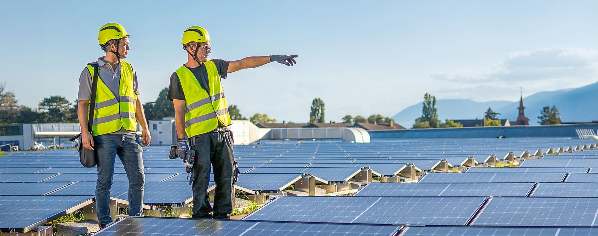 Zwei Arbeiter in Warnwesten und Schutzhelmen stehen auf einem Dach mit Photovoltaik-Anlagen; einer zeigt in die Ferne, während der andere ihm aufmerksam zuhört.