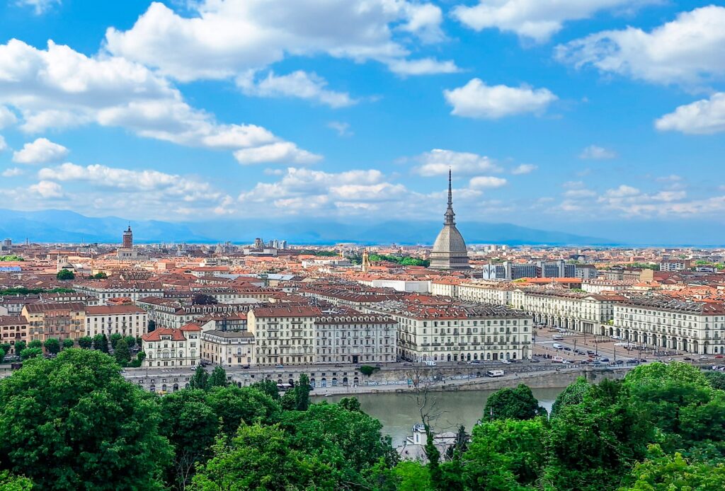 Das Panoramabild zeigt eine weitläufige Stadtansicht unter einem blauen Himmel mit vielen weissen Wolken. Im Zentrum dominiert die markante Kuppel der Mole Antonelliana die Skyline. Dicht bebaute Häuserreihen erstrecken sich bis zum Horizont, wo im Hintergrund sanfte Berge sichtbar sind, während im Vordergrund ein Fluss und üppige grüne Bäume die Szene abrunden.