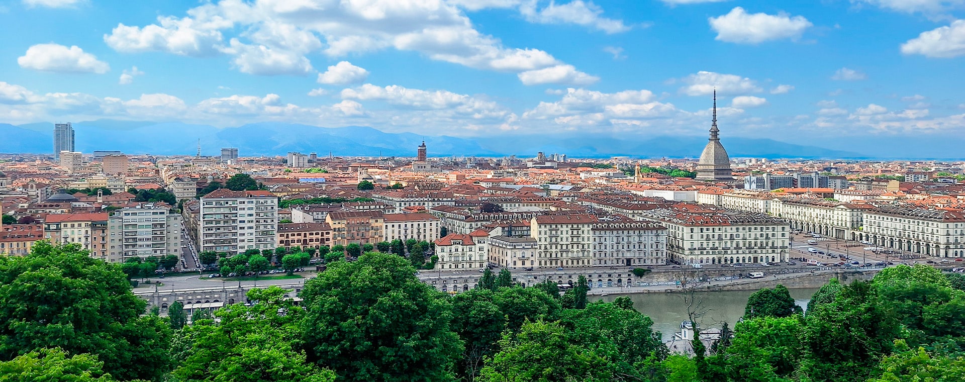 Das Panoramabild zeigt eine weitläufige Stadtansicht unter einem blauen Himmel mit vielen weissen Wolken. Im Zentrum dominiert die markante Kuppel der Mole Antonelliana die Skyline. Dicht bebaute Häuserreihen erstrecken sich bis zum Horizont, wo im Hintergrund sanfte Berge sichtbar sind, während im Vordergrund ein Fluss und üppige grüne Bäume die Szene abrunden.